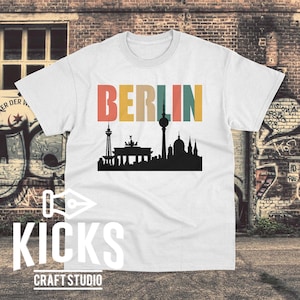 May include: White t-shirt with the word "BERLIN" in colorful block letters above a black silhouette of Berlin landmarks. The shirt also features the text "KICKS CRAFT STUDIO" in white.