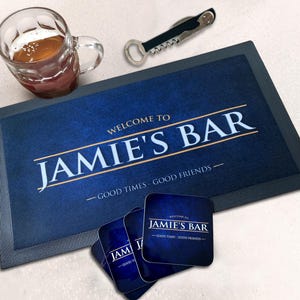 Może przedstawiać: Prostokątna ciemnoniebieska mata barowa i trzy kwadratowe podstawki z napisem "WELCOME TO JAMIE'S BAR" oraz "GOOD TIMES - GOOD FRIENDS" w białym i złotym kolorze, w zestawie ze szklanym kuflem do piwa i metalowym otwieraczem do butelek.