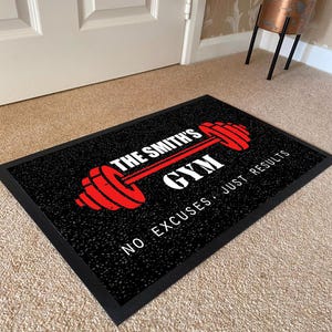 May include: A rectangular black doormat with a textured surface featuring a red barbell graphic and white text that reads THE SMITH'S GYM and NO EXCUSES JUST RESULTS. The mat has a solid black border and is positioned on a light-colored carpet.