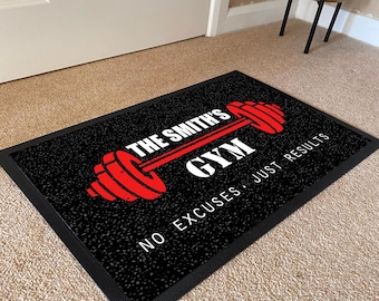 May include: A rectangular black doormat with a textured surface featuring a red barbell graphic. The text reads THE SMITH'S GYM in white and red, with the phrase NO EXCUSES · JUST RESULTS printed in white below the barbell graphic.