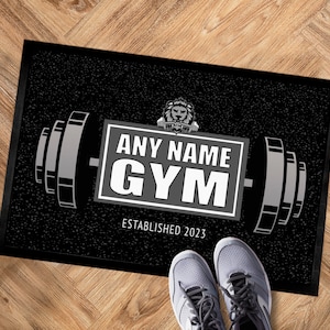 Pode incluir: Tapete preto e branco com um design de barra e o texto "ANY NAME GYM ESTABLISHED 2023". Um logotipo de cabeça de leão está acima do texto.
