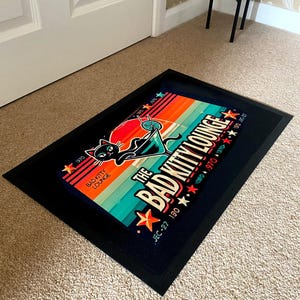 May include: A rectangular black doormat with a colorful graphic. The graphic features a black cat in a martini glass, the text "The Bad Kitty Lounge", and star accents. The background has orange, teal, and blue stripes.