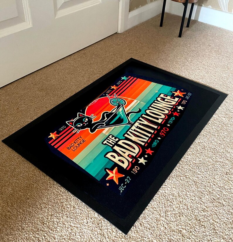 May include: A rectangular black doormat with a colorful graphic. The graphic features a black cat in a martini glass, the text "The Bad Kitty Lounge", and star accents. The background has orange, teal, and blue stripes.