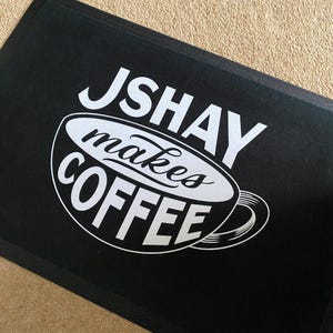 May include: Black rectangular doormat with the text "JSHAY makes COFFEE" in white. The text is arranged around a stylized coffee cup design. The mat is made of a durable material.