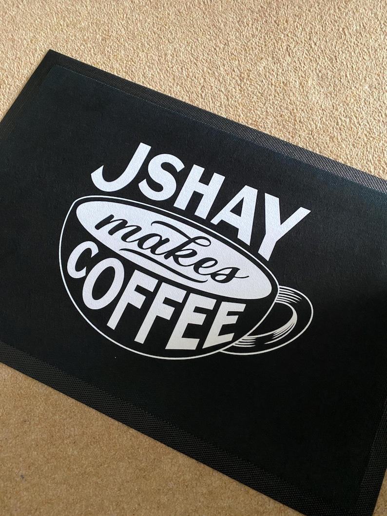 May include: Black rectangular doormat with the text "JSHAY makes COFFEE" in white. The text is arranged around a stylized coffee cup design. The mat is made of a durable material.