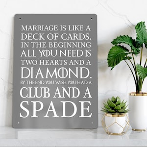 Könnte beinhalten: Ein rechteckiges graues Metallschild mit weißem Text: MARRIAGE IS LIKE A DECK OF CARDS. IN THE BEGINNING ALL YOU NEED IS TWO HEARTS AND A DIAMOND. BY THE END YOU WISH YOU HAD A CLUB AND A SPADE. Das Schild hat kleine Löcher in den Ecken.