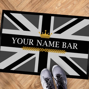 Peut inclure: Un paillasson noir avec un motif de drapeau Union Jack gris et blanc. Le drapeau a une couronne dorée et le texte "YOUR NAME BAR" en blanc.