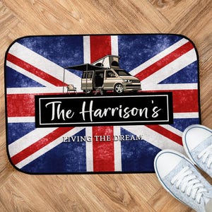 May include: Rectangular doormat featuring a distressed Union Jack design with a camper van illustration. The text "The Harrison's" is displayed in a black rectangle, with "LIVING THE DREAM" below. A pair of light blue sneakers are visible.