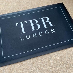 May include: A rectangular black doormat with the text "TBR LONDON" in white. The doormat has a white border and a textured black edge. The mat is placed on a light brown carpet.