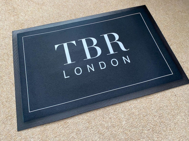 May include: A rectangular black doormat with the text "TBR LONDON" in white. The doormat has a white border and a textured black edge. The mat is placed on a light brown carpet.