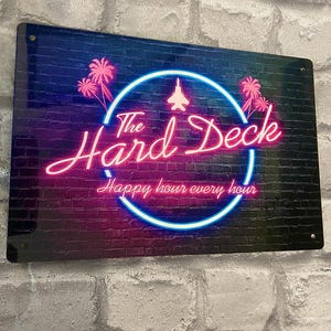 Puede incluir: Un letrero rectangular con un diseño de neón. El letrero presenta las palabras "The Hard Deck" en una fuente cursiva, con un círculo azul neón y un gráfico de chorro. El letrero también incluye el texto "Happy hour every hour."