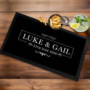 Peut inclure: Un panneau rectangulaire noir avec les noms "LUKE & GAIL" et la date "27 juin 2026" en texte blanc. Le panneau est encadré de noir. Un bol de collations, un cocktail et un verre à shot sont sur la surface en bois.