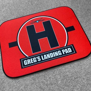 Peut inclure: Tapis d'atterrissage pour drone rectangulaire rouge avec une bordure noire. Le tapis présente un grand "H" noir à l'intérieur d'un cercle blanc, un graphique de drone et le texte "GREG'S LANDING PAD" dans un rectangle blanc.