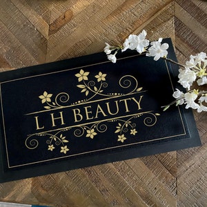 May include: Black rectangular mat with gold floral design and the text "LH BEAUTY". The mat is on a wooden surface, with white flowers in the upper right corner. The mat has a gold border.