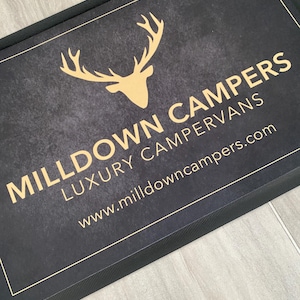 May include: A rectangular black doormat with a gold-colored deer head logo and text that reads "MILLDOWN CAMPERS LUXURY CAMPERVANS" and "www.milldowncampers.com". The mat has a textured surface and a black border.