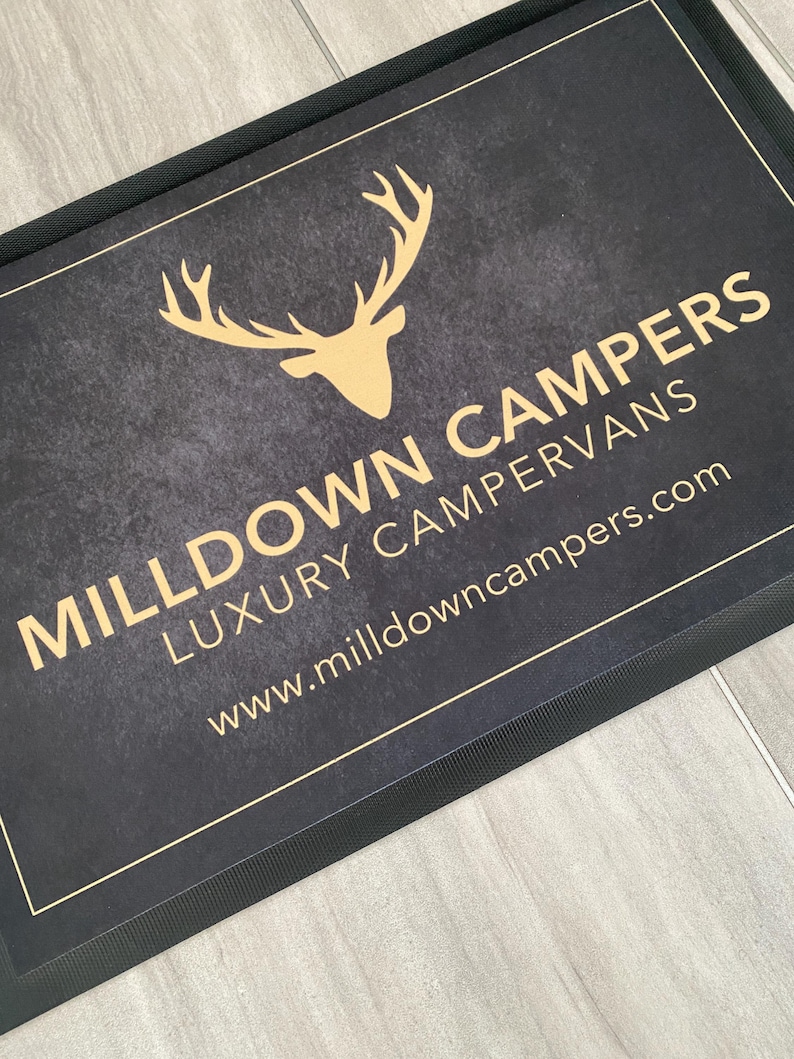 May include: A rectangular black doormat with a gold-colored deer head logo and text that reads "MILLDOWN CAMPERS LUXURY CAMPERVANS" and "www.milldowncampers.com". The mat has a textured surface and a black border.