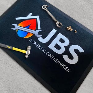 May include: Black rectangular mat with the JBS Domestic Gas Services logo, a house icon, and a wrench. A small hammer, wrench, and copper fittings are on the mat.