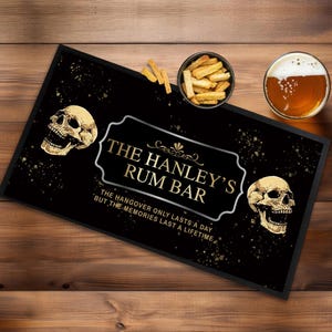 Op de afbeelding: Zwarte barmat met gouden glitter en schedels. De tekst luidt "The Hanley's Rum Bar. The hangover only lasts a day, but the memories last a lifetime."