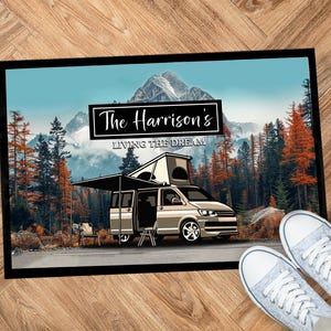 May include: A rectangular doormat with a black border features a camper van parked on a road in front of a mountain range and forest. The text "The Harrison's LIVING THE DREAM" is displayed above the van. A pair of light blue trainers are in the foreground.