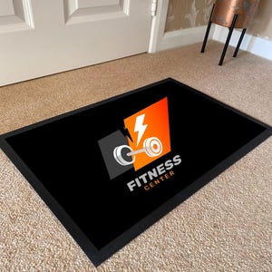 May include: Black rectangular doormat with a black border. The mat features a logo with a barbell, lightning bolt, and the words "FITNESS CENTER" in white. The logo is set against a gray and orange background.