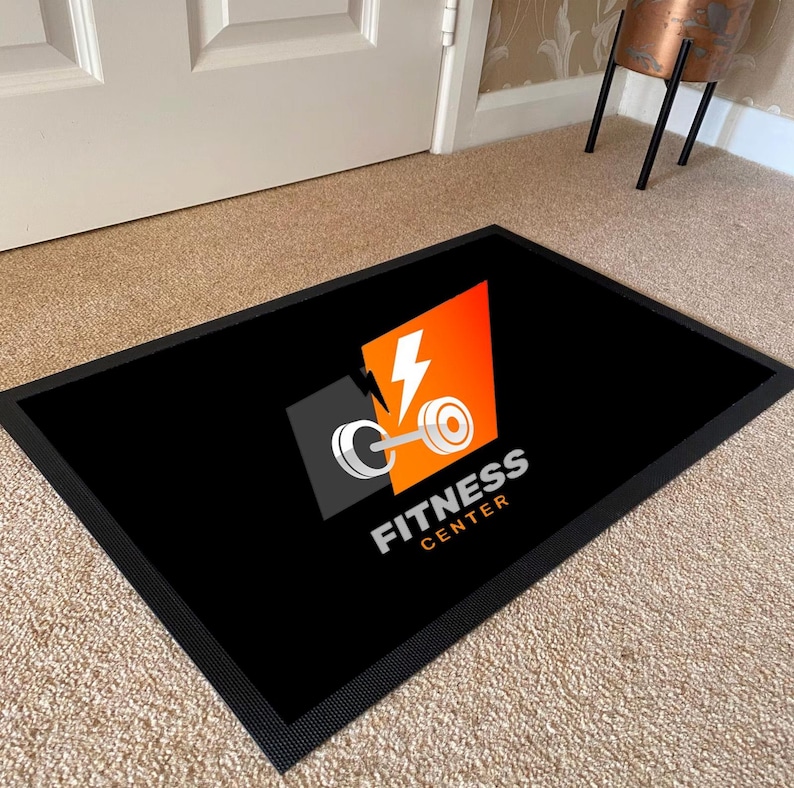 May include: Black rectangular doormat with a black border. The mat features a logo with a barbell, lightning bolt, and the words "FITNESS CENTER" in white. The logo is set against a gray and orange background.