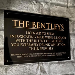 Peut inclure: Panneau rectangulaire noir avec texte doré. Il indique "THE BENTLEYS" et précise que l'établissement est autorisé à servir des boissons enivrantes. Il comprend également une clause de non-responsabilité concernant la perte de la parole, du contrôle de soi ou des vêtements.