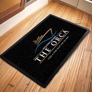Op de afbeelding: Zwarte rechthoekige deurmat met een zwarte rand. De mat heeft een gouden en blauwe afbeelding van een boot met de witte woorden "THE ORCA". Daaronder staat de zin "TIME & TIDE WAIT FOR NO ONE."