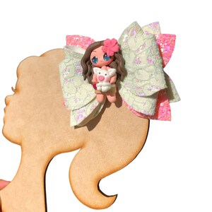 Puede incluir: Un lazo verde y rosa con un dise&ntilde;o de encaje blanco. Una peque&ntilde;a mu&ntilde;eca con cabello casta&ntilde;o y un cerdo est&aacute; en el centro del lazo.
