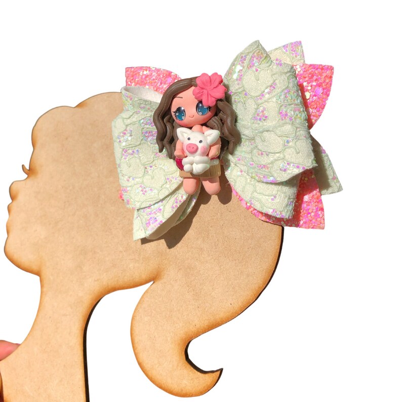 Puede incluir: Un lazo verde y rosa con un dise&ntilde;o de encaje blanco. Una peque&ntilde;a mu&ntilde;eca con cabello casta&ntilde;o y un cerdo est&aacute; en el centro del lazo.