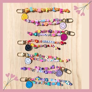 May include: A collection of colorful beaded keychains with various words and smiley face charms. The keychains feature words like "SIMPLE", "TEEEWS", "LEAH", "PRETTY", "LOVELY", "CUTE", and "SMILE". Each keychain has a gold-colored clasp.