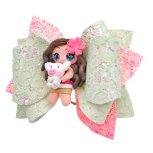 Puede incluir: Un lazo de brillo verde y rosa con una figura de mu&ntilde;eca hecha a mano que sostiene un cerdo. La mu&ntilde;eca tiene cabello casta&ntilde;o, ojos azules y lleva un vestido rosa y una falda marr&oacute;n.