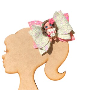 Puede incluir: Un lazo rosa y blanco con un acabado brillante presenta una peque&ntilde;a mu&ntilde;eca con un cerdo. La mu&ntilde;eca tiene el pelo casta&ntilde;o, una flor rosa y ojos grandes. El lazo est&aacute; sujeto a una cabeza de mu&ntilde;eca de madera.