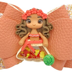 Puede incluir: Un lazo marr&oacute;n y naranja con una peque&ntilde;a mu&ntilde;eca de pelo casta&ntilde;o que lleva una corona de flores rojas y un vestido rojo y blanco. La mu&ntilde;eca sostiene un amuleto redondo verde con un dise&ntilde;o.