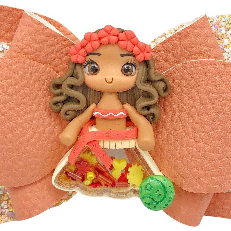 Puede incluir: Un lazo marr&oacute;n y naranja con una peque&ntilde;a mu&ntilde;eca de pelo casta&ntilde;o que lleva una corona de flores rojas y un vestido rojo y blanco. La mu&ntilde;eca sostiene un amuleto redondo verde con un dise&ntilde;o.