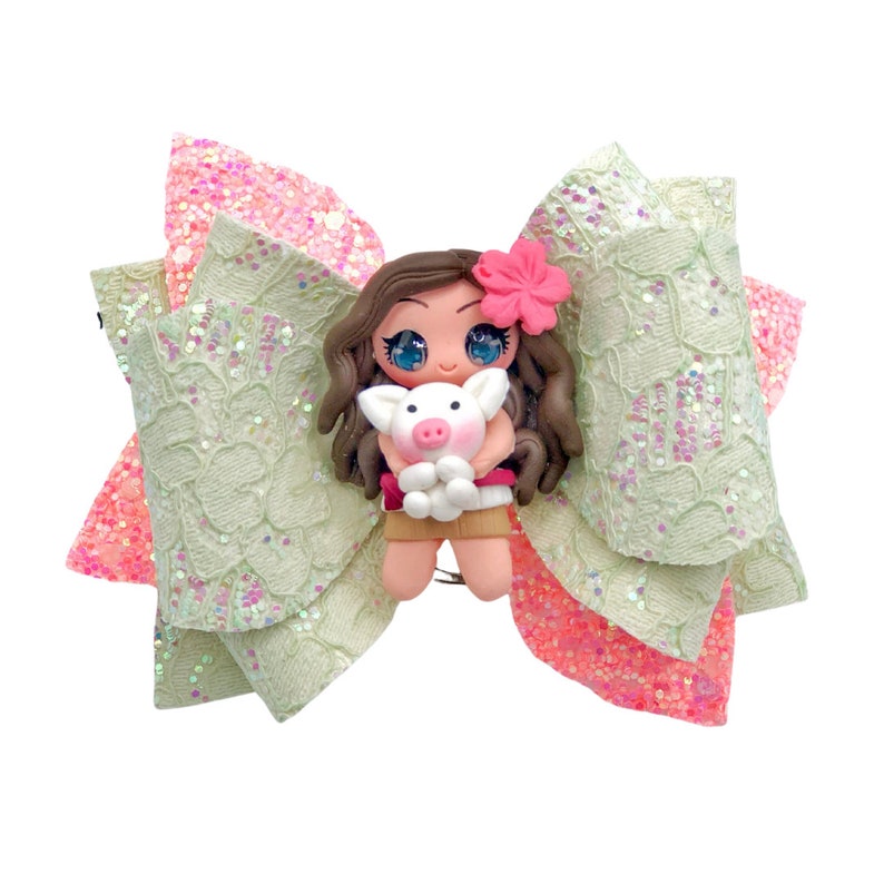 Puede incluir: Un lazo verde y rosa con una peque&ntilde;a mu&ntilde;eca que sostiene un cerdo. La mu&ntilde;eca tiene el pelo casta&ntilde;o, una flor rosa y un vestido rosa y blanco. El lazo est&aacute; hecho de encaje y purpurina.