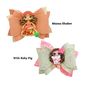 Puede incluir: Dos lazos para el pelo hechos a mano con una mu&ntilde;eca de Moana. El primer lazo es de color melocot&oacute;n con purpurina y presenta a Moana sosteniendo un coco. El segundo lazo es verde y rosa con purpurina y presenta a Moana sosteniendo un peque&ntilde;o cerdo. El texto "Moana Shaker" y "With Baby Pig" es visible en la imagen.