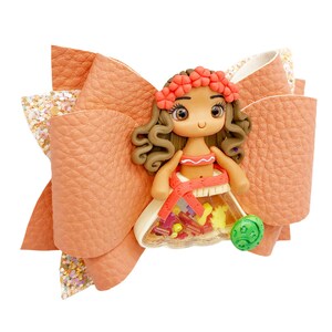 Puede incluir: Un lazo de tela color melocot&oacute;n con un acento de brillo y una peque&ntilde;a figura de mu&ntilde;eca en el centro. La mu&ntilde;eca lleva un atuendo rojo y naranja con una corona de flores y un amuleto verde.