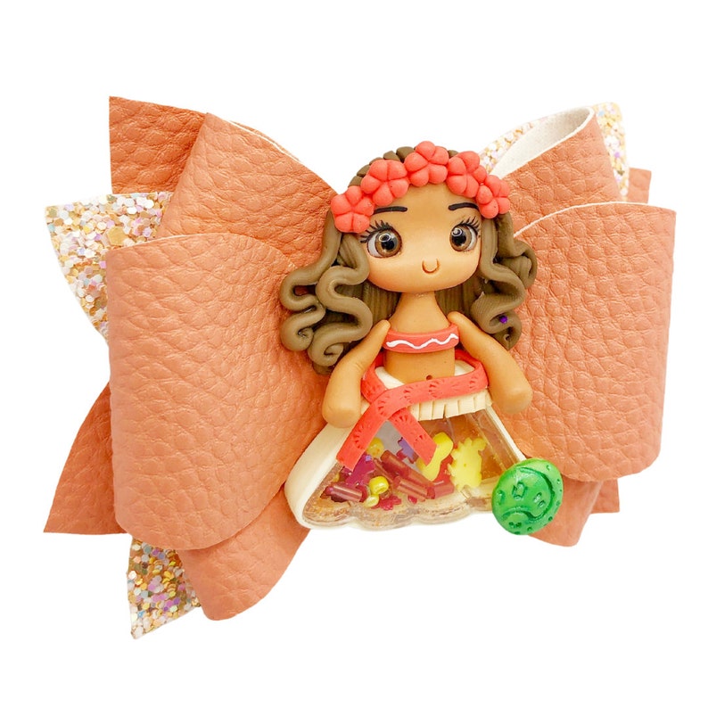 Puede incluir: Un lazo de tela color melocot&oacute;n con un acento de brillo y una peque&ntilde;a figura de mu&ntilde;eca en el centro. La mu&ntilde;eca lleva un atuendo rojo y naranja con una corona de flores y un amuleto verde.