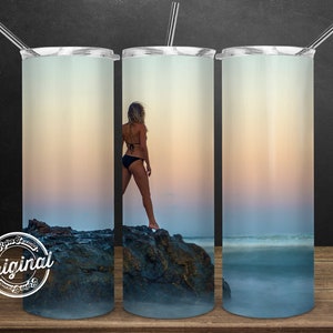 May include: A stainless steel tumbler with a colorful sunset scene and a woman standing on a rocky shore. The tumbler has a clear lid and a straw.