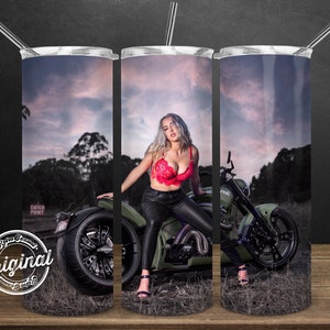 May include: A stainless steel tumbler with a colorful image of a woman sitting on a green motorcycle. The background is a sunset sky with clouds. The tumbler has a straw and a lid.