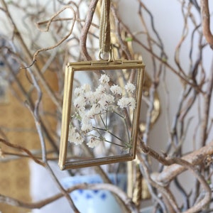 May include: A gold-toned picture frame with a delicate white flower pressed inside. The frame is hanging from a branch with a brown velvet ribbon.