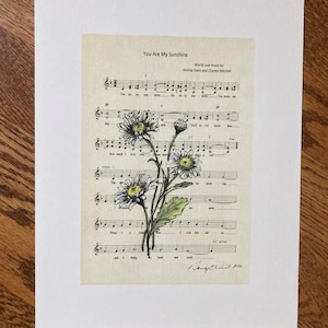 Puede incluir: Una impresión de partitura vintage de la canción "You Are My Sunshine" con margaritas dibujadas a mano en primer plano. La partitura incluye la letra y las notas musicales. Las palabras "Words and music by Jimmie Davis and Charles Mitchell" están impresas en la parte superior de la partitura.