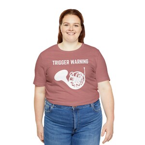 Puede incluir: Una mujer con una camiseta color marrón rojizo con el texto "TRIGGER WARNING" y una silueta blanca de un trompa.