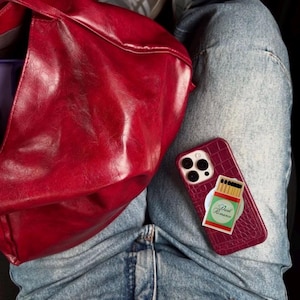 May include: A burgundy leather tote bag and a matching phone case with a matchbox design are visible. The phone case has a textured finish and a pop-socket with the words "Dark Romance". The person is wearing blue jeans.