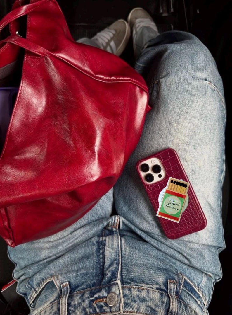 May include: A burgundy leather tote bag and a matching phone case with a matchbox design are visible. The phone case has a textured finish and a pop-socket with the words "Dark Romance". The person is wearing blue jeans.