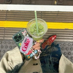 May include: A clear plastic cup filled with a green beverage and a green straw. A phone with a decorative case and a Diet Coke can are held together. The background is a train platform.