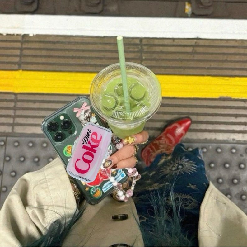 May include: A clear plastic cup filled with a green beverage and a green straw. A phone with a decorative case and a Diet Coke can are held together. The background is a train platform.