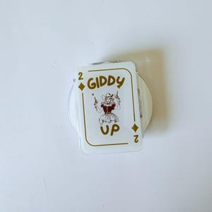May include: A white playing card with gold text and a cartoon image of a cowgirl holding two guns. The card reads "2 GIDDY UP" in gold lettering. The card is a novelty item.