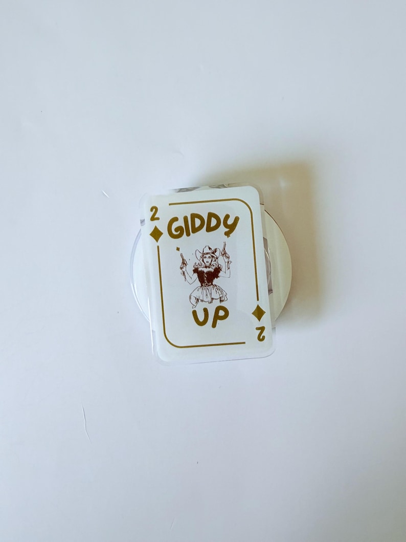May include: A white playing card with gold text and a cartoon image of a cowgirl holding two guns. The card reads "2 GIDDY UP" in gold lettering. The card is a novelty item.