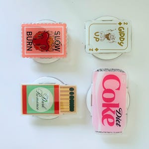 May include: Four decorative phone grips with unique designs. One features a flame with "SLOW BURN" text, another a playing card with "2 GIDDY UP", a third with "Dark Romance" and matches, and a pink Diet Coke can.